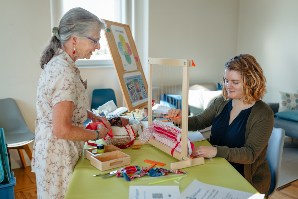One woman sitting and working on a weaving art project with another woman standing and watching her