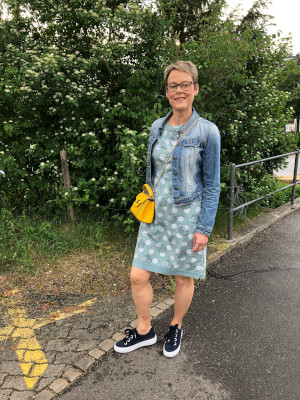 The picture shows Rachel, who received support from Cancer Support Switzerland, She is standing outside on a cement sidewalk with trees behind her.